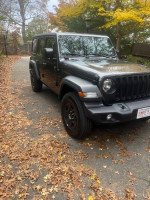 2023 Jeep Wrangler Sport 4WD in grey parked on a leaf-covered path, showcasing its rugged design.
