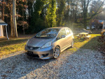 2010 Honda Fit Sport in silver parked in a gravel area surrounded by trees, highlighting its hatchback design and 96,000 miles.
