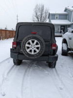 Rear view of a 2014 Jeep Wrangler Sahara 4WD in snow, showcasing its tires and black exterior.