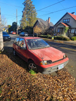 1990 Geo Metro parked on the roadside, showing wear and leaves scattered around it.