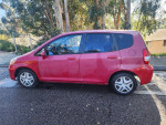 Side view of a 2007 Honda Fit in red, showcasing its compact design and manual transmission features.