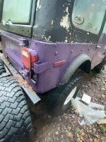 Rear view of a purple 1978 Jeep CJ7 with rugged tires, showing wear and tear, parked in a wooded area.